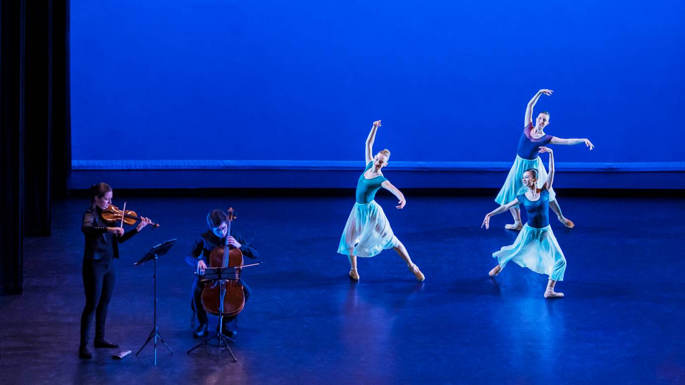 Three women in flowing skirts extend a leg back with their arms in high third; two musicians play